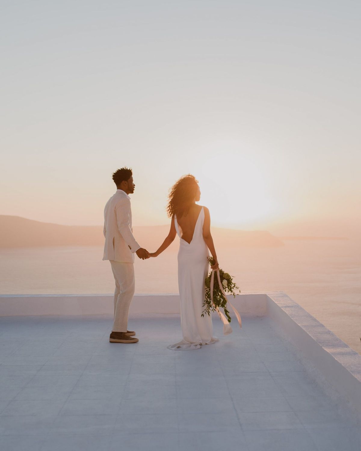 poročne fotografije sončnega zahoda neveste in ženina, ki se držijo za roke na strehi v Grčiji